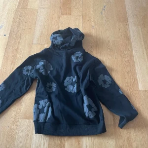 Svart hoodie med gråa blommor Denim Tears - Svart hoodie från Denim Tears med stora gråa blommönster över hela tröjan. Tröjan har huva och är långärmad, perfekt för dig som gillar streetwear och vill sticka ut med en unik design. Tillverkad i mjuk bomull för skön känsla.