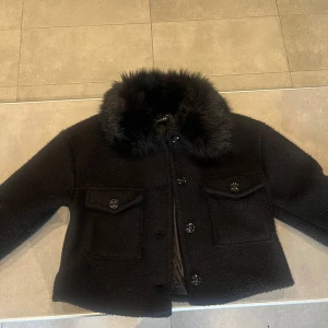 Svart teddyjacka med pälsig krage - Snygg svart teddyjacka med fluffig pälsimitation på kragen. Jackan har två stora fickor framtill, knäppning med knappar och en croppad passform. Perfekt för dig som vill ha en meotine liknande jacka