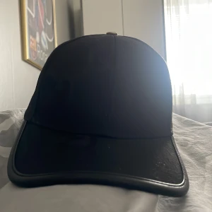 Svart keps från Gucci - Snygg svart keps från Gucci med klassisk böjd skärm och justerbart spänne bak i läder. Kepsen är tillverkad i Italien och har diskret Gucci-logga på baksidan. Perfekt accessoar för en clean och lyxig look. Helt ny köpte den men sen jag tänkte sälja den igen för mindre pris går bak på det men ja tänkte köpa en annan nu 