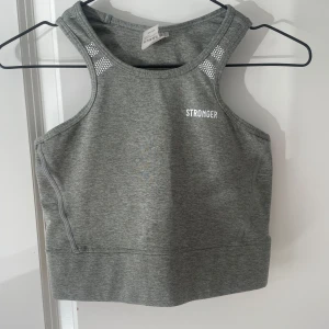 Grå linnetopp från Stronger - Sportig grå linnetopp från Stronger med vita detaljer och logga framtill. Toppen har brottarrygg och små silvriga prickar vid axlarna. Tillverkad i stretchigt material som andas, perfekt för träning och aktiva dagar. Knappt använd. Finns matchande byxor till i en annons