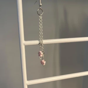 Silverfärgat örhänge med rosa blommor - Hängande örhänge i silverfärg med två tunna kedjor och små ljusrosa blommor längst ner. Söt och trendig detalj som ger en lekfull touch till din stil. Det är handgjorda!