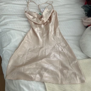 Champagnefärgad satin klänning  - Supersnygg kort klänning i champagnefärgad satin från Gina tricot. Klänningen har tunna axelband, draperad urringning och öppen rygg med snygga snörningsdetaljer. 