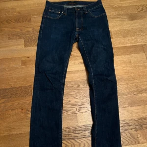 Mörkblå jeans från Nudie Jeans - Säljer ett par mörkblå jeans från Nudie Jeans med klassisk femficksdesign och kontrastsömmar. Jeansen har raka ben och snygg broderad detalj på bakfickan samt läderpatch i midjan. Perfekta för dig som gillar stilrena och tidlösa jeans.