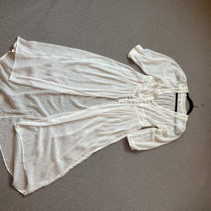Vit vadlång klänning/tunika Vintage Stories - Romantisk vit långklänning från Vintage Stories by Kappahl i transparent, prickigt tyg med spetsdetaljer. Klänningen har v-ringning, knappar framtill och trekvartsärmar. Perfekt för dig som gillar vintage vibes och flowy siluett.