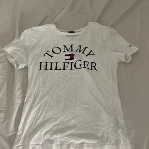 Vit t-shirt från Tommy Hilfiger - Vit t-shirt från Tommy Hilfiger med stor logga och flagga framtill. Klassisk rund hals och korta ärmar. Tillverkad i ekologisk bomull för en skön och mjuk känsla. Perfekt basic-plagg med snygg passform och stilren look.