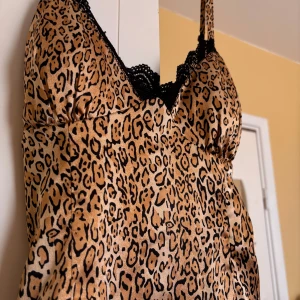 Leopardmönstrad linnetopp från Guess - Snygg leopardmönstrad linnetopp från Guess med tunna axelband och svart spetsdetalj vid urringningen. Toppen är i glansigt stretchigt material som känns lyxigt och har en figurnära passform. Perfekt för dig som vill sticka ut med en djurisk vibe.