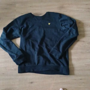 Svart tröja från Lyle & Scott - Svart sweatshirt från Lyle & Scott med klassisk rund hals och gul broderad logga på bröstet. Tröjan har långa ärmar och ribbade muddar vid ärmslut och nederkant. Perfekt för en chill och stilren look. Pris kan diskuteras 