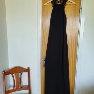 Svart långklänning med spetsdetaljer - Elegant svart långklänning med spets vid axlarna och öppen rygg. Klänningen har en slits där det har spruckit i sömmen men ytterst lite. Kolla bilderna. Klänningen är armlös med hög halsringning. Perfekt för dig som vill ha en stilren och sofistikerad look. Perfekt till baler! 