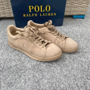 Beige mockasneakers från Polo Ralph Lauren - Stilrena sneakers i beige mocka från Polo Ralph Lauren. Klassisk låg modell med rund tå, platt sula och snörning. Diskret logga broderad på sidan och Polo-tryck på sulan. Perfekta för en clean och avslappnad look.