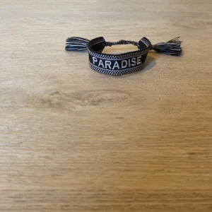 Svart och vit armband med text - Snyggt vävt armband i svart och vitt med texten 'PARADISE' broderad framtill. Armbandet har justerbar knytning med tofsar i ändarna och ett coolt, grafiskt mönster längs kanterna. Perfekt accessoar för att lyfta din outfit.  