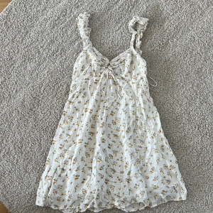 Vit blommig kortklänning Bardot - Supersöt vit kortklänning från Bardot med små gula blommor, volangiga axelband och snörning framtill. Lätt och flowy modell, 