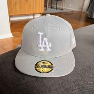 Grå LA-keps New Era 59FIFTY 7 1/8 - Snygg grå keps från New Era, modell 59FIFTY med LA-logga broderad framtill i vitt. Klassisk flat peak och broderad New Era-logga på sidan. Kepsen är i bomull och har en stilren look som passar perfekt till streetwear.