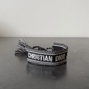 Svart armband från Christian Dior - Snyggt armband från Christian Dior i svart och vitt med tydlig logga framtill. Armbandet har broderade detaljer och avslutas med tofsar. Perfekt accessoar för att lyfta din outfit.