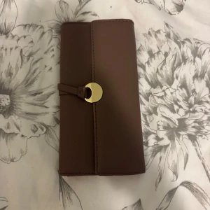 Brun plånbok med gulddetalj - Säljer en stilren brun plånbok med snygg guldfärgad knapp framtill. Plånboken har flera fack för kort och ett fönster för ID. Perfekt för att hålla ordning på dina kort och kontanter.