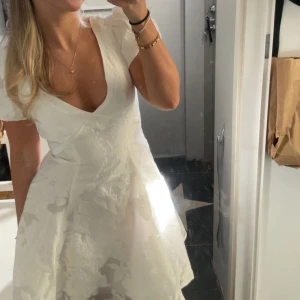 Studentklänning - Superfin vit klänning med v-ringning och puffiga kortärmar. Klänningen har en lätt utställd kjol och ett subtilt blommigt mönster i tyget. Perfekt för sommarens tillfällen och studentfirande. Materialet känns lyxigt och har struktur. Sömmen på ena axeln har sprättats upp lite (de bild) men det är något som enkelt går att sy! ☀️Nypris: 1087kr