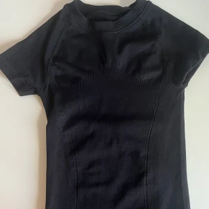 Svart tight träningstopp - Svart kortärmad träningstopp med tight passform och diskreta ribbade detaljer. Perfekt för gymmet eller löprundan. Materialet är stretchigt och följsamt för maximal rörelsefrihet.