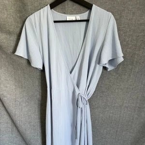 Ljusblå omlottklänning från Vila - Superfin ljusblå omlottklänning från Vila med korta, vida ärmar och knyt i midjan. Klänningen har en v-ringad hals och är tillverkad i ett mjukt, lätt material som passar perfekt till varmare dagar.