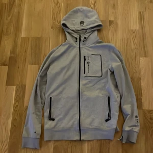 Ljusgrå hoodie från Sail Racing - Ljusgrå hoodie med dragkedja från Sail Racing. Jackan har huva, svarta dragkedjor på fickor och bröstficka samt tryckdetaljer på ärm och bröst. Perfekt för en sportig och avslappnad stil.