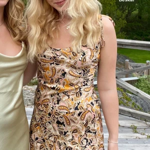 Paisleymönstrad klänning från Neon Rose - Klänning från Neon Rose i satinliknande material. Klänningen har smala axelband och en figurnära passform. Färgerna går i rosa, orange, svart och beige.