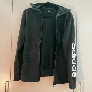 Svart hoodie med dragkedja från Adidas - Svart hoodie från Adidas med vit logga längs ena ärmen och liten logotyp på ryggen. Tröjan har huva, dragkedja och två fickor framtill. Perfekt för en sportig och avslappnad stil. Storleken är 38. Priset kan diskuteras. 
