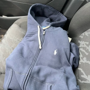Mörkblå hoodie från Ralph Lauren - Mörkblå hoodie med dragkedja från Ralph Lauren. Klassisk design med vit broderad logga på bröstet och vit dragsko i huvan. Perfekt för en avslappnad stil.