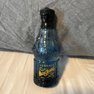 Versace Blue Jeans Man Eau de Toilette - Versace Blue Jeans Man är en herrparfym i en blå glasflaska med unikt mönster och gul text. Flaskan har en klassisk, nästan retro design med detaljerade präglingar och svart kork med grekiskt mönster. Det är en 75 ml flaska som är använd några gånger men finns fortfarande stor del kvar av den 