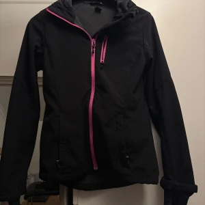 Svart vindjacka från H&M med rosa dragkedja - Svart vindjacka från H&M med snygga rosa dragkedjor. Jackan har långa ärmar och en sportig look, perfekt för blåsiga dagar. Justerbar nederkant och praktiska fickor framtill. Enkel och stilren design.