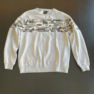 Grå sweatshirt med camouflagemönster  - Grå sweatshirt med en bred panel i camouflagemönster över bröstet och axlarna som sträcker sig runt hela kroppen. Najs passform, storlek:M. 