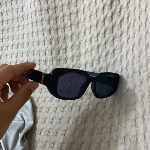 Svarta solglasögon från Yves Saint Laurent - Snygga svarta solglasögon från Yves Saint Laurent med rektangulära bågar och guldiga YSL-detaljer på skalmarna. Glasögonen har en modern oversized look och är tillverkade i Italien.