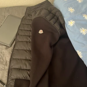 Svart pufferjacka från Moncler - Säljer en svart pufferjacka från Moncler med quiltade partier och ribbade muddar. Jackan har lång ärm och Moncler-logga på ena ärmen. Perfekt för kyligare dagar och riktigt snygg till streetwear-stil.