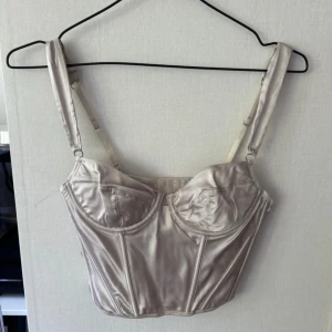 Champagnefärgad satin korsettopp - Snygg champagnefärgad korsettopp i satin med justerbara axelband och markerade kupor. Toppen har en croppad passform och dekorativa sömmar framtill för en trendig look. Litet slitage som man ser på bild 2, dock endast använd 2 gånger.