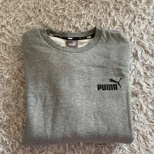 Grå sweatshirt från Puma - Säljer en klassisk grå sweatshirt från Puma med svart logga på bröstet. Tröjan har rund halsringning och långa ärmar. Mjuk insida och ribbade muddar vid ärmslut och nederkant. Perfekt för en avslappnad stil. Inga defekter. 