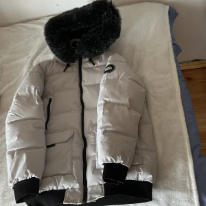 Vit pufferjacka med svart fuskpäls från Supply & Demand - Säljer en vit pufferjacka från Supply & Demand med svart fuskpäls på huvan. Jackan har svarta detaljer, dragkedja framtill, fickor med lock och ribbade muddar. Perfekt för kalla dagar och riktigt snygg streetstil.