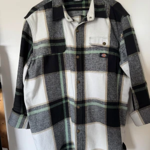 Rutig overshirt från Dickies - Säljer en snygg rutig overshirt från Dickies i svart, grått, vitt och grönt. Skjortan har långa ärmar, bröstficka med knapp och klassisk krage. Perfekt att slänga över en t-shirt för en avslappnad stil.