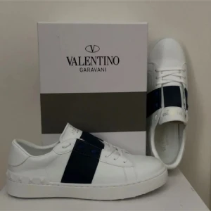 Valentino Garavani vita sneakers med svart detalj - Snygga Valentino Garavani sneakers i vitt med en bred blå rand över ovansidan. Skorna har en klassisk design med snörning och en diskret logotyp i guld på tungan. Tyvär inte legit.Perfekta för en stilren look. Strl 44. Nyskick. Säljer pga reselling. Öpen till prisförslag 