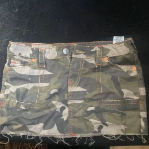 Kjol true religon - Säljer en cool camouflage kortkjol från true religon med råa kanter och praktiska fickor. Kjolen har orange sömmar som ger en extra touch. Perfekt för en avslappnad stil.
