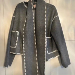 Grå kappa från Nelly - Grå kappa från Nelly med dekorativa vita sicksack-kanter längs krage, fickor och ärmslut. Kappan har stora framfickor och knäppning framtill. Rak och något oversized passform, perfekt för lager-på-lager. Knappt använd, köpt för 700kr. Storlek S men skulle säga passar xs och xxs också.