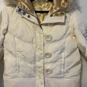 Vit pufferjacka från DKNY Active med fuskpäls - Säljer en vit pufferjacka från DKNY Active med stor huva och fuskpäls. Jackan har stora knappar framtill, dragkedja och ribbade muddar. Snygg detalj med DKNY-logga och nitar på ärmen samt glansigt innerfoder.