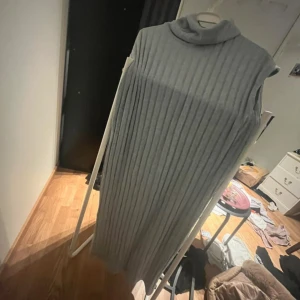 Ljusgrå ribbad långklänning från Lindex - Säljer en ljusgrå långklänning från Lindex med polokrage och ribbad struktur. Klänningen är ärmlös och har en rak, loose passform som ger en avslappnad look. Perfekt för lager-på-lager-stil under kallare dagar.