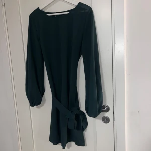 Mörkgrön klänning med knytband - Säljer en stilren mörkgrön klänning med långa ärmar och ett brett knytband i midjan. Klänningen har en rak passform och är perfekt för dig som gillar enkel men elegant stil