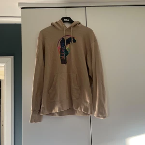 Beige hoodie från Uniqlo - Säljer en beige hoodie från Uniqlo i samarbete med Musée du Louvre och Peter Saville. Tröjan har en färgglad konstinspirerad print framtill och huva. Knappt använd. Stilren.
