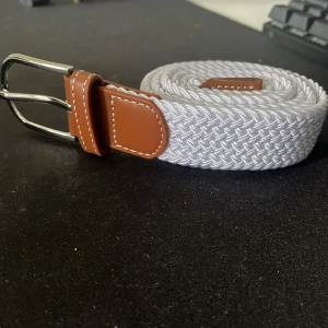 Vitt flätat bälte med bruna skinn-detaljer  - Snyggt vitt flätat bälte med detaljer i brunt skinn och silvrigt spänne. Bältet har en klassisk design med vävd struktur och passar perfekt till jeans eller chinos. Enkel att justera tack vare det flexibla materialet.