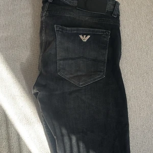 Svarta jeans från Armani med slitningar - Strl 32, Svarta jeans från Armani med tydliga slitningar och hål på flera ställen. Klassisk femficksmodell med broderad logga på bakfickan. Jeansen har normal passform och är tillverkade i denim.