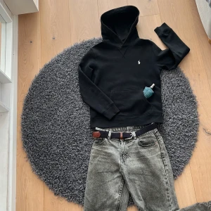 Svart hoodie från Ralph Lauren - Säljer en stilren svart hoodie från Ralph Lauren med en liten broderad logga på bröstet. Tröjan har en klassisk känguruficka och en bekväm huva. Perfekt för en avslappnad stil.