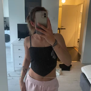 Svart croppad linnetopp med vita ränder - Säljer en svart croppad linnetopp med tunna vita vertikala ränder och smala axelband. Toppen har en asymmetrisk nederkant och knytning i sidan för extra detalj.