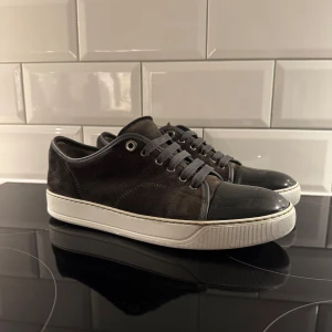 Grå lanvins  - Säljer ett par mörkgrå sneakers från Lanvin med vit, tjock sula och svarta skosnören. Skorna har rund tå och detaljer i både mocka och skinn, samt metallhål för snörningen. Perfekta för en stilren och avslappnad look.