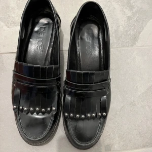 Svarta loafers med fransar och nitar från Bianco - Säljer ett par svarta loafers från Bianco i glansigt skinn. Skorna har dekorativa fransar och silvriga nitar framtill. Klassisk rund tå och låg platt sula. Perfekta för dig som vill ha en stilren men edgy look.