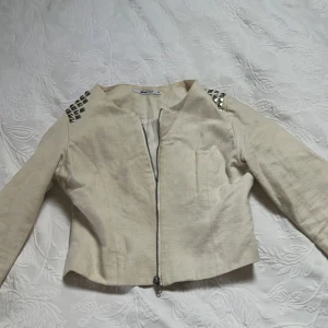 Beige jacka med nitar på axlarna - Säljer en beige jacka med coola silvriga nitar på axlarna och sned dragkedja framtill. Jackan är croppad och har lång ärm, perfekt för dig som vill sticka ut lite extra. Snygg och trendig modell som passar till många outfits.