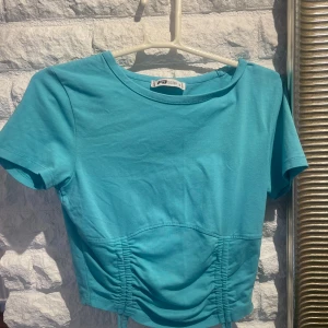 Turkos croppad topp med rynkade detaljer - Säljer en turkos croppad topp från Gina Tricot med korta ärmar och snygga rynkade detaljer framtill. Perfekt för sommardagar och enkel att matcha med jeans eller kjol.
