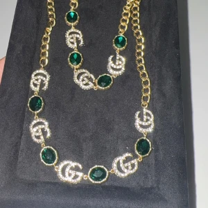 GG halsband och armband med gröna stenar - Säljer ett exklusivt set från GG med halsband och armband i guldfärgad metall, dekorerade med gröna stenar och glittrande GG-loggor. Kedjorna är robusta och detaljerade, perfekt för att lyfta din outfit. 